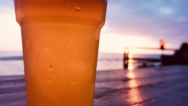 A glass of beer is sitting on a table by the ocean. The glass is half full and the beer is cold