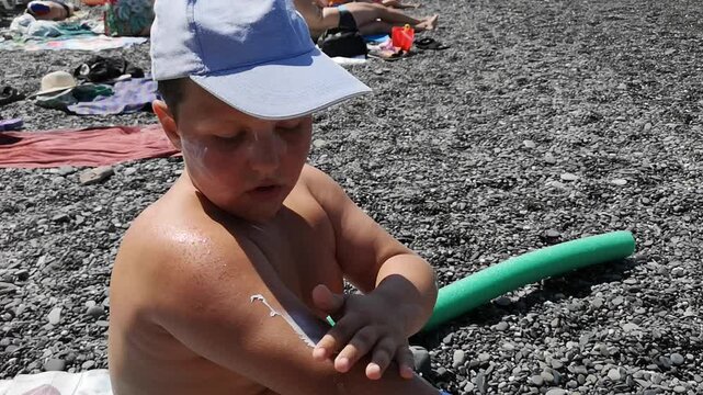 A boy rubs sunscreen onto his skin. He sits on a pebble.