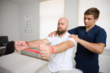 Physiotherapist guiding male patient with resistance band exercise for shoulder rehabilitation in clinic. Physical therapy session focused on injury recovery, strengthening, mobility improvement