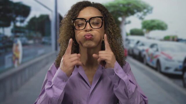 Transgender hispanic woman with glasses presses fingertips to puffed cheeks amid blurred parked cars on street; playfulness.