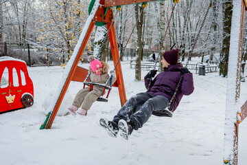 A father with his daughter, a three-year-old girl, on a swing on a snowy playground in winter