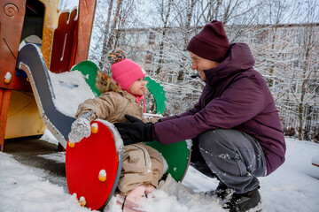 A father and his daughter, a three-year-old girl, play on the playground in winter, dad catches the child from the slide