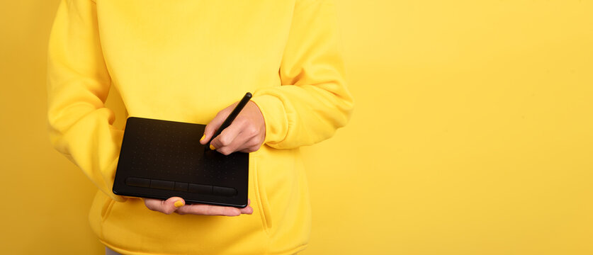 Person in yellow sweater holding and drawing on a black graphic tablet against a solid yellow background. Modern technology and creativity concept.