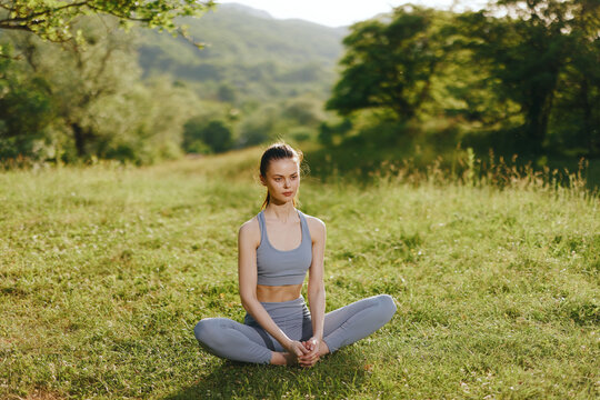 Serene young woman practicing yoga outdoors in a green landscape, wearing gray athletic clothing and exuding tranquility, suitable for wellness and fitness themes