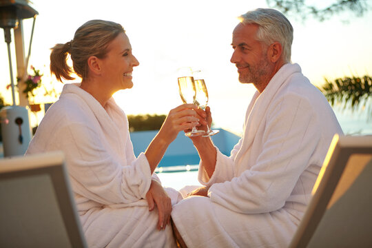Sunset, robe and couple in resort with cheers, bonding or anniversary celebration on spa trip. Honeymoon, man or woman poolside with champagne toast, connection or summer evening in wellness getaway.