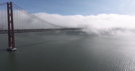 Aerial drone shot towards the 25th April Bridge and waterfront in Lisbon, Portugal, Europe. Sunny and bright with clouds, fog, mist covering the bridge. Shot in ProRes 422 HQ