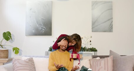 Diverse couple at home with woman covering man's eyes, man unwrapping and peeking into gift box - Powered by Adobe