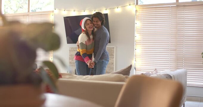 Couple wearing Santa hat grasping hands and dancing under string lights at home for holiday