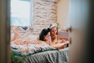 Two women spending quality time at home, wearing headbands and eye masks, relaxing on the bed while focusing on a tablet screen. The scene radiates friendship, closeness, and home comfort.