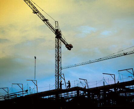 Silhouette of workers and cranes at a construction site during sunset, showing industrial development and urban growth. Modern building framework with safety nets and structural scaffolding.