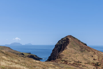 Dry volcanic hill at Ponta Sao Lourenco Madeira overlooking distant islands, narrow summit trail with hikers rises above calm blue Atlantic water