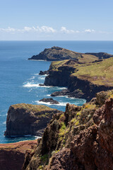 Vertical postcard view of Ponta de Sao Lourenco Madeira with full rocky headland and distant lighthouse, revealing layered cliffs above bright blue water