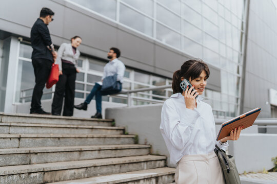 A motivated businesswoman talks on a smartphone and works on a tablet outside an office building, while colleagues interact in the background, portraying a dynamic and communicative business setting.