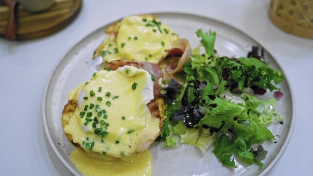 Eggs benedict with creamy hollandaise sauce and fresh salad served on a white plate in a cafeteria setting showcasing a delicious breakfast meal.