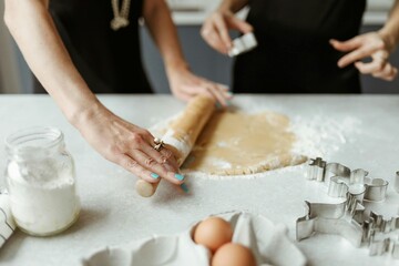 Close-up of hands rolling out homemade cookie dough with a wooden rolling pin on a floured kitchen counter, surrounded by baking ingredients like eggs, flour, and metal cookie cutters, illustrating