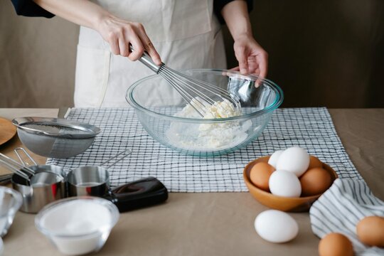 Person in a beige apron whisking white ingredients in a clear glass mixing bowl, surrounded by baking essentials like eggs, a sieve, measuring cups, and a checkered towel, preparing homemade dessert.