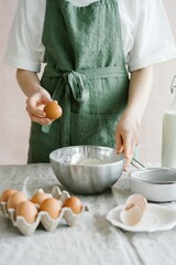 Person in green apron cracking egg into a bowl of flour, with fresh ingredients like eggs, milk, and baking tools on a rustic linen tablecloth, showcasing the process of homemade baking or cooking.