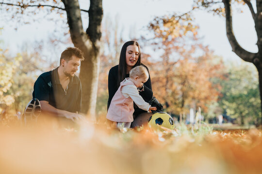 A family enjoys a sunny autumn day in the park. A mother and father with their baby explore the grass, near a yellow soccer ball, surrounded by colorful fall foliage.