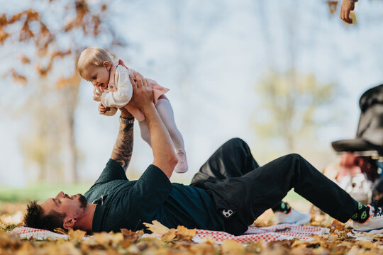 A joyful father rests on a checkered blanket in a sunlit autumn park, lifting his baby daughter into the air. Warm light, fallen leaves, and a tender family moment outdoors.