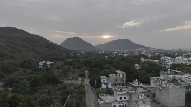 Aerial shot of Jamini Kund Road in Pushkar during the evening, showcasing a warm sunset behind the Aravali hills with a dramatic cloudy sky and a clear view of the Pushkar townscape below.