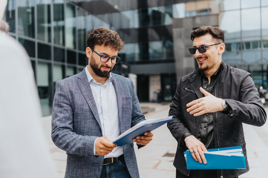 Two business colleagues standing outdoors reviewing documents and conversing next to an office complex.