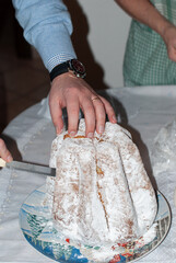 A hand cuts into a tall, sugar-dusted holiday cake on a festive table, capturing a warm, cozy moment of traditional Christmas dessert preparation.