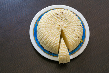 Вelicious round white homemade cheese with textured, ridged surface and slice cut out, placed on decorative plate with blue and gold rim, revealing its creamy interior