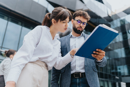 Two business colleagues are analyzing documents outside a modern office building, collaborating efficiently to discuss strategies and plans.