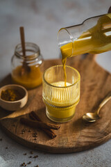 Ein Frau f&uuml;llt Goldene Milch in ein Trinkglas auf einem grauen Tisch. Heissgetr&auml;nk, Winter.