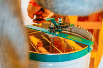 Close-up of a beekeeping centrifugal honey extractor in action. Green metal gears and frame mechanism over a stainless steel drum. A hand operates the crank, extracting honey from frames inside the dr