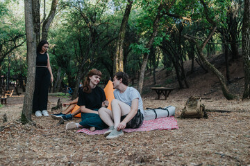 A couple sits on a checkered blanket in a shaded woodland, chatting and smiling while a friend stands nearby. Camping gear and a backpack rest beside them for a casual outdoor retreat.