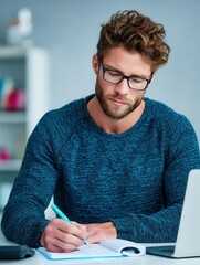 A young man with glasses is deeply focused on writing in his notebook at a modern workspace, conveying concentration and productivity.