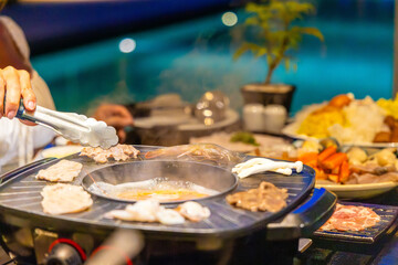 Asian hot pot dinner, seafood and vegetables on grill, hand using tongs, outdoor dining, vibrant colors, social gathering, evening meal.