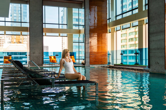 Woman in white swimsuit relaxing by indoor hotel pool with cityscape background. Urban leisure, luxury travel, resort lifestyle.