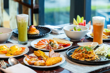 Diverse breakfast spread with fresh juice on restaurant table. Includes eggs benedict, cold cuts, noodles, tropical fruits. Concept of brunch, variety, dining out.