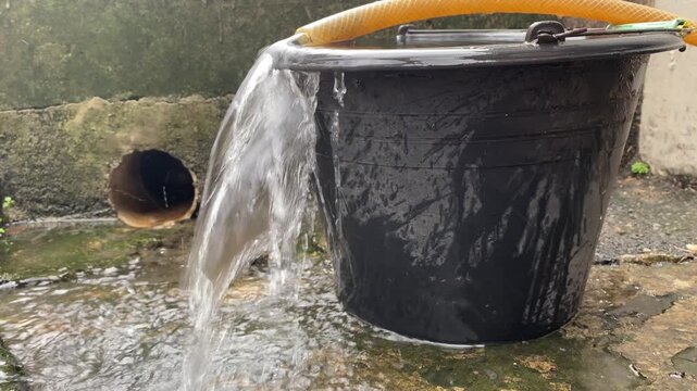 Clear water flows from a hose into a black bucket. Water is poured into a bucket. In the barrel water is collected from a hose