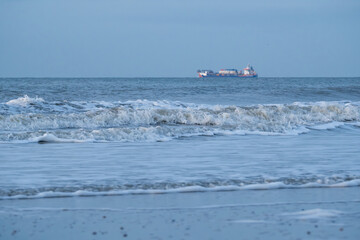 Waves crashing on a serene beach with a cargo ship in the distance under a soft blue sky during twilight