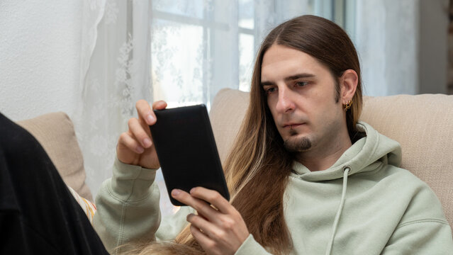 Young man with long hair relaxing on a couch, focused on reading an e-reader in a cozy indoor setting