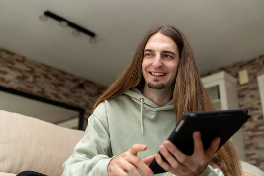 Young man with long hair and hoodie sitting on a couch, smiling while browsing content on his tablet, enjoying technology
