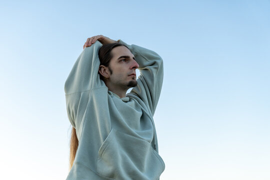 Man with long hair stretching arms above head against clear blue sky, finding peace and relaxation outdoors