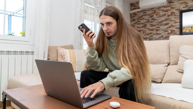 Young man with long hair multitasking at home, working on laptop while talking on smartphone from living room sofa - Powered by Adobe
