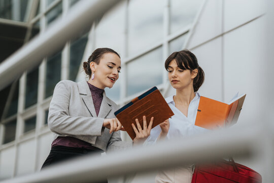 Two motivated business professionals reviewing documents and discussing strategies outdoors near a corporate structure.