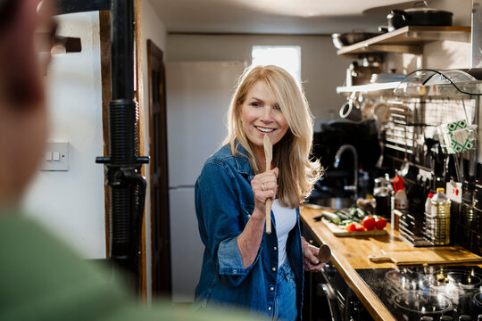 Woman in a kitchen smiling and holding a wooden spoon playfully