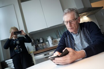 Man using smartphone at breakfast table while woman takes photo in kitchen