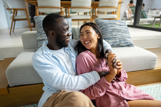 Excited child-free multiethnic couple embracing and laughing in modern living room, calm lifestyle - Powered by Adobe