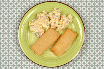 An overhead shot of two breaded fish fillets and French salad (Croatian way) served on a bright green plate against a patterned tablecloth