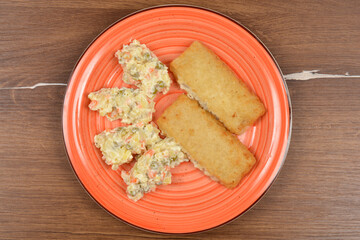 An overhead view of two breaded fish filets and several dollops of French salad (Croatian way) on a vibrant orange plate set against a dark wood surface