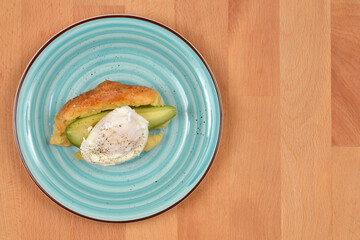A croissant with a poached egg and avocado is served on a striped blue plate placed on a light wooden surface