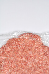 A close-up view captures a raw ground pork patty sealed in clear plastic wrap against a white background