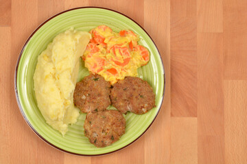 A green plate contains three meat patties, a portion of mashed potatoes, and sliced carrots in a creamy sauce, set on a wooden table